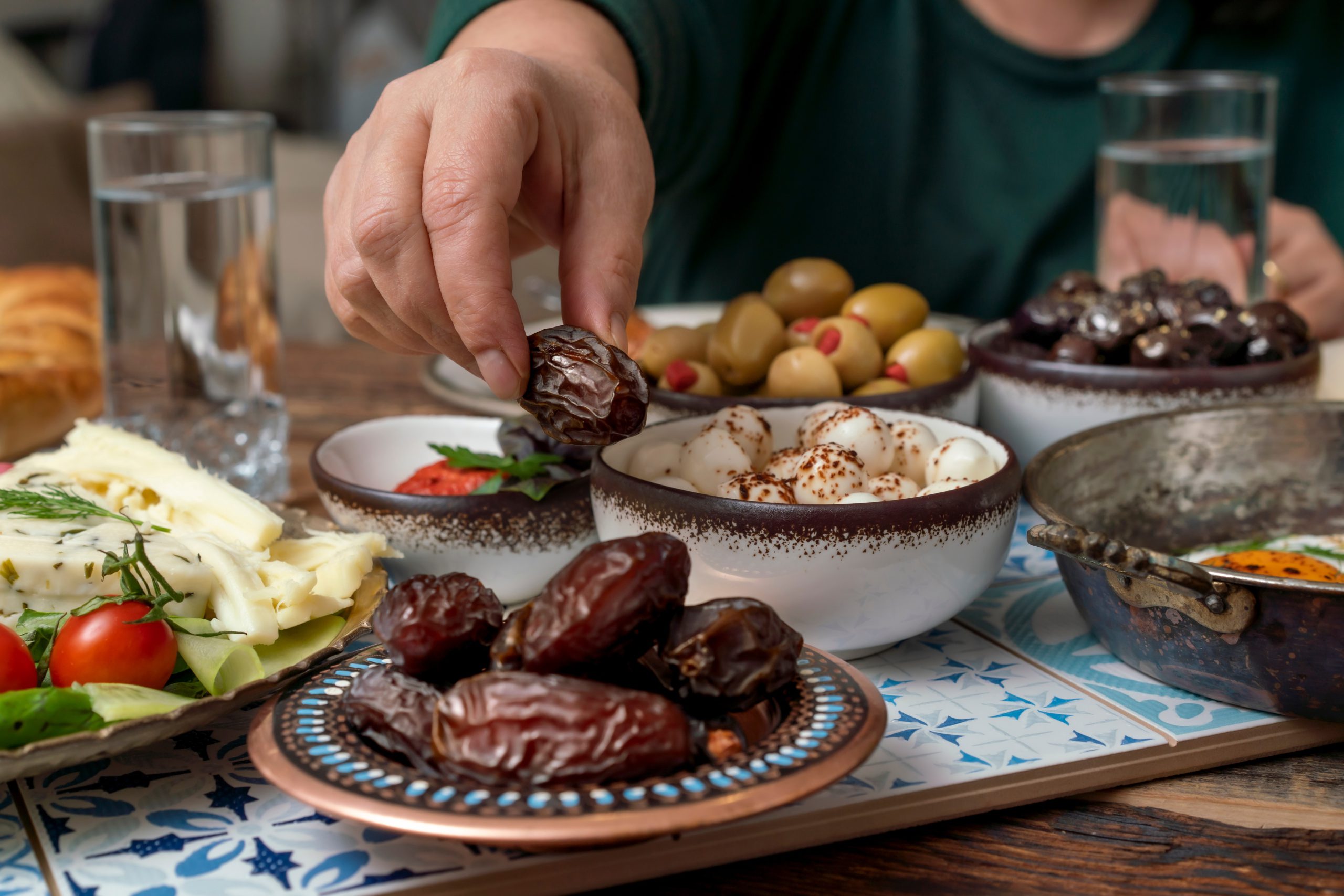 Person picking up a date from a traditional iftar meal spread, featuring dates, olives, cheeses, vegetables, and glasses of water